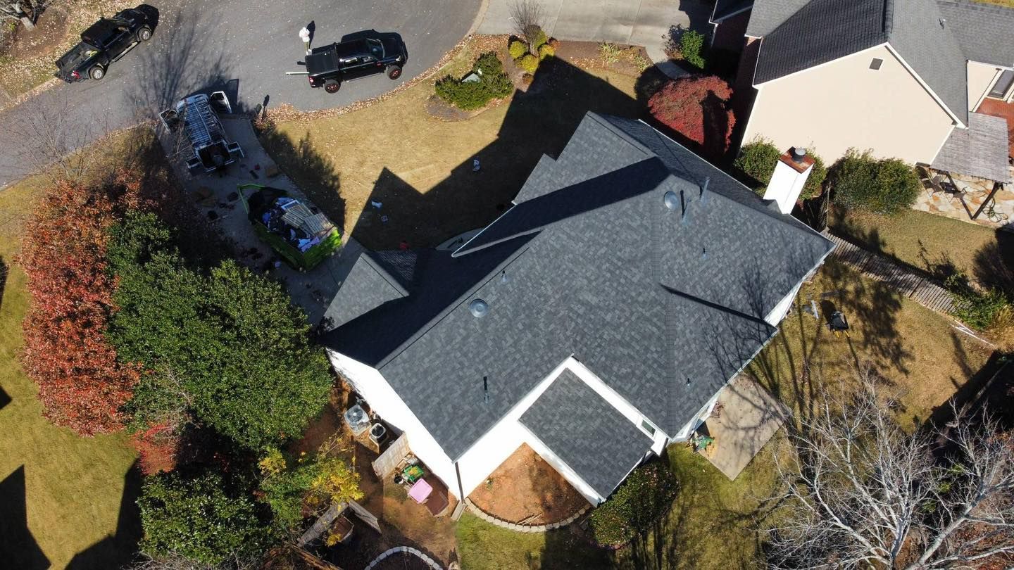 Aerial view of a house with a dark gray roof, surrounded by lawn and trees. Driveway with cars.