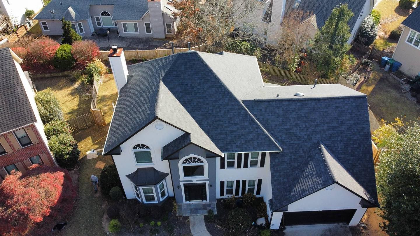 Aerial view of a gray-roofed house with white siding and black shutters, set in a neighborhood with trees.