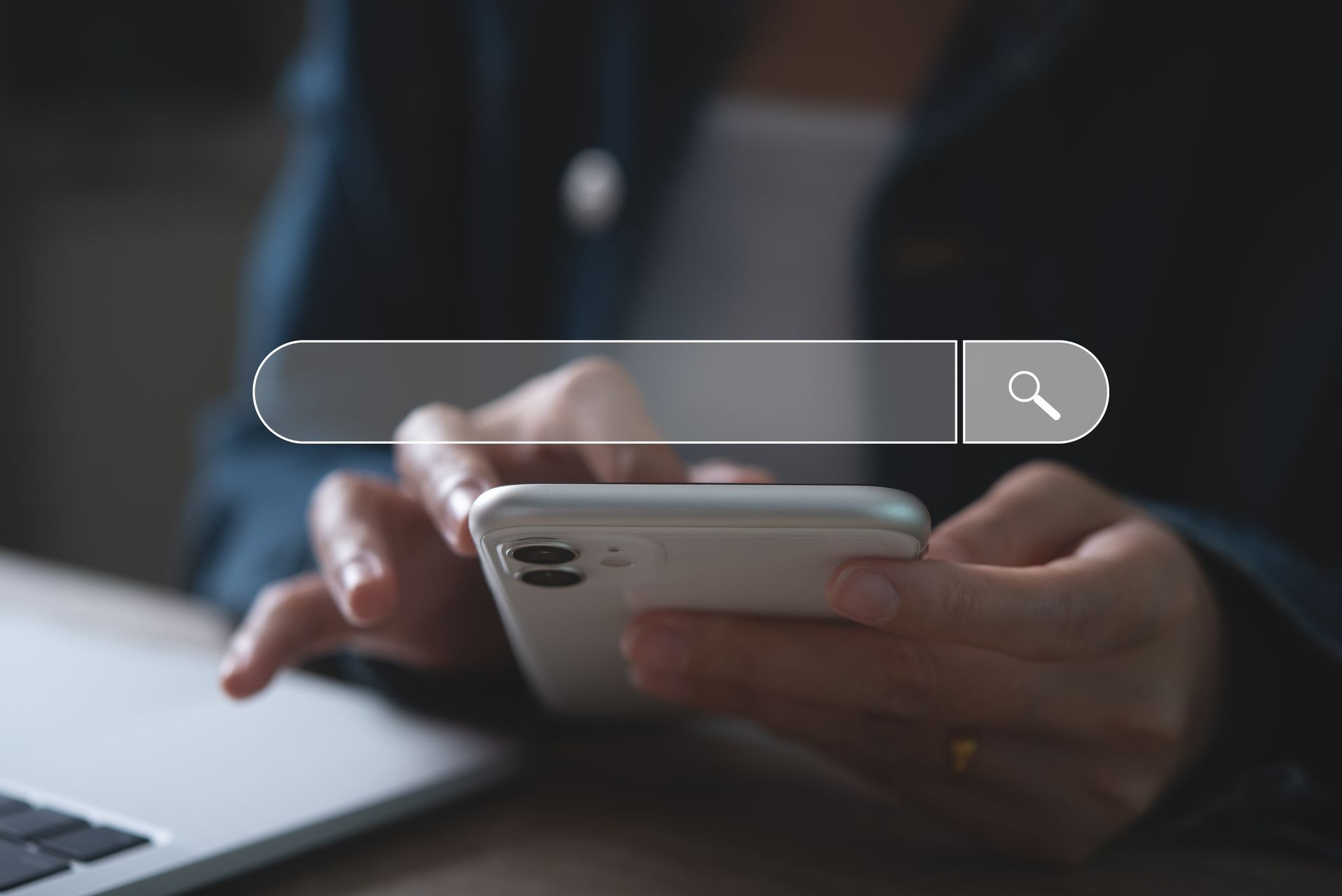 Person typing on a smartphone beside a laptop, with a search bar overlay on the screen.