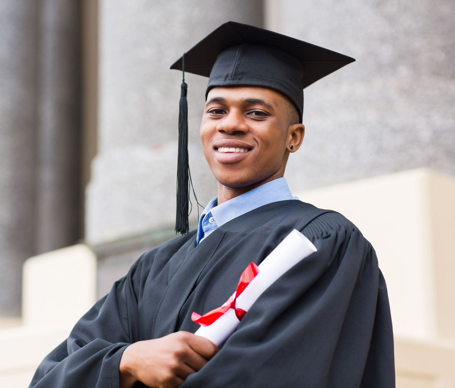 Graduate in cap and gown holding a diploma, smiling outdoors on steps.