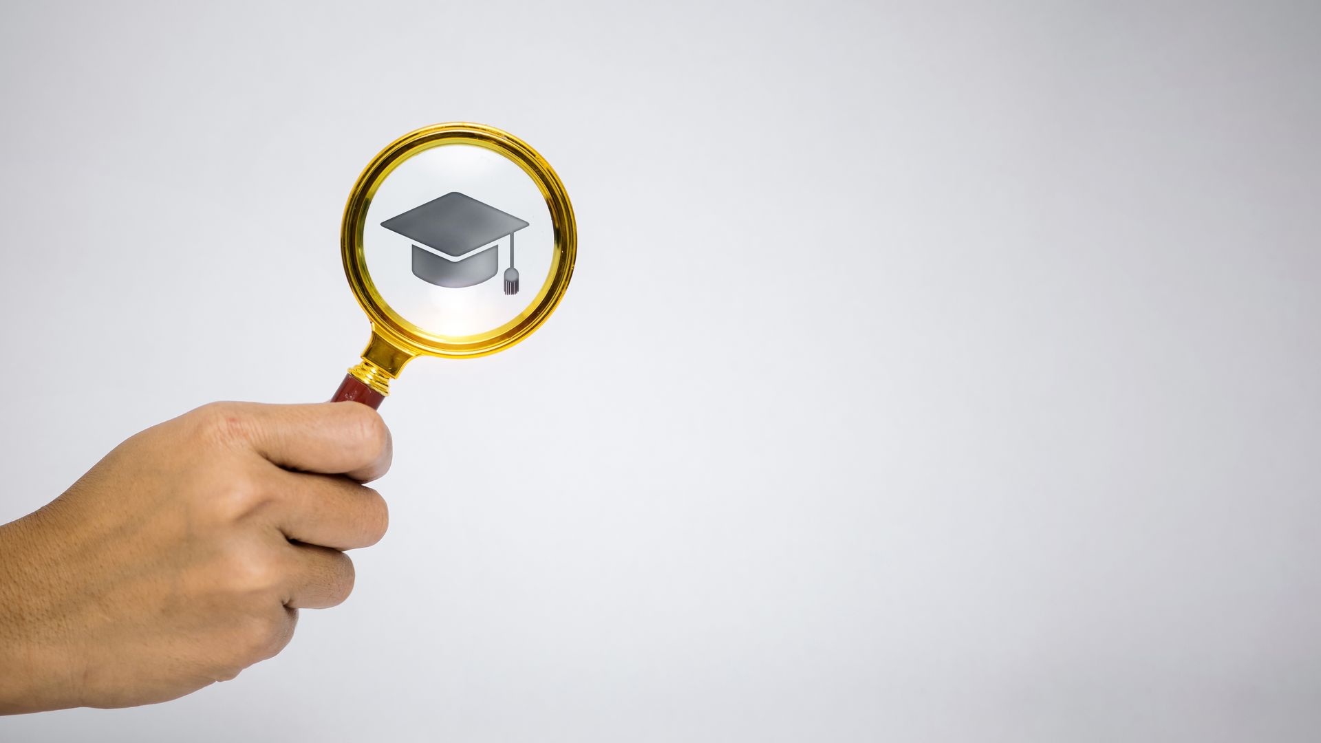 Hand holding a gold magnifying glass over a graduation cap icon on a light background