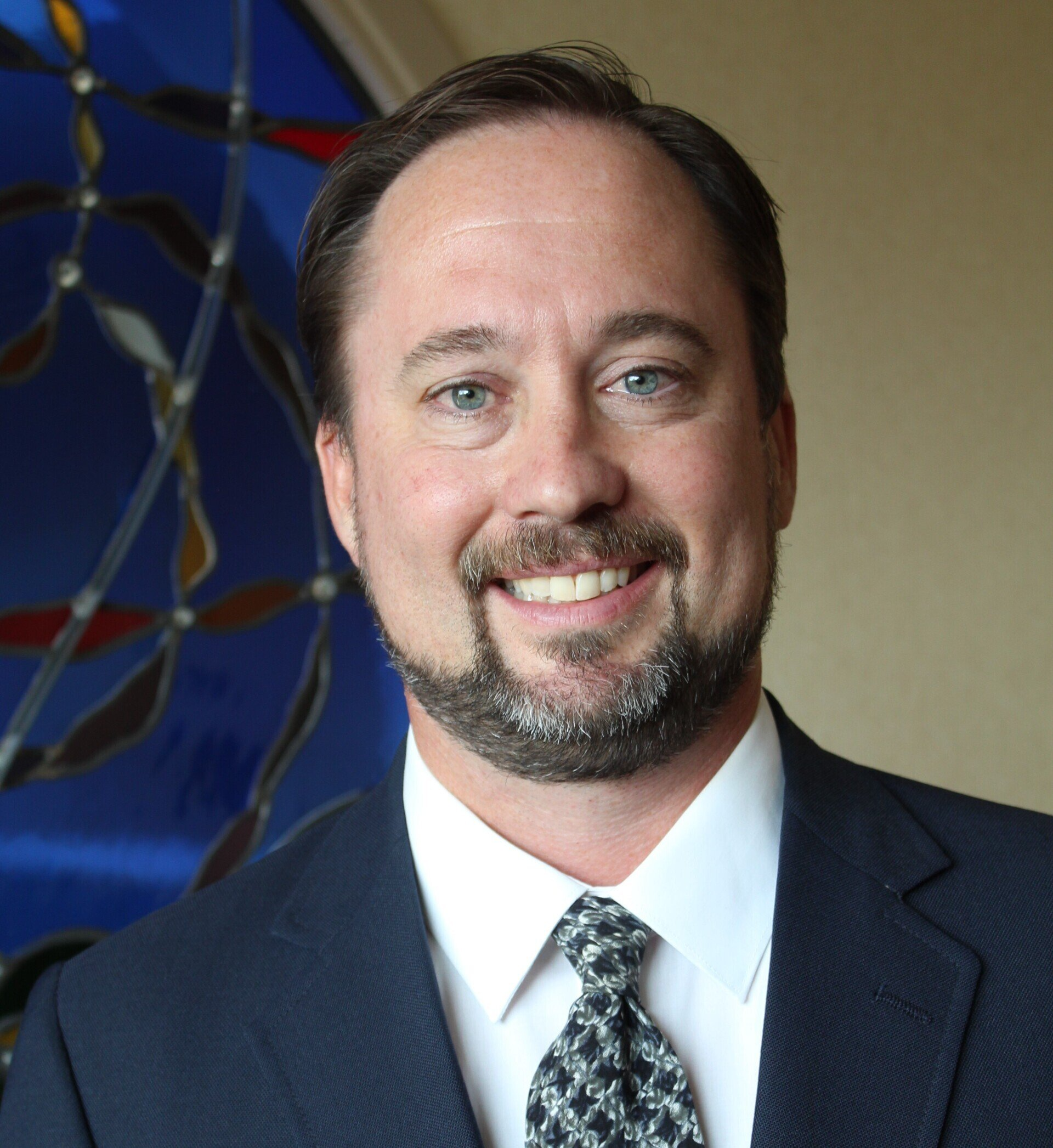 Man in suit smiles, standing near a stained glass window.