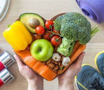 Hands holding a heart-shaped bowl filled with colorful fruits and vegetables, fitness equipment visible in the background.