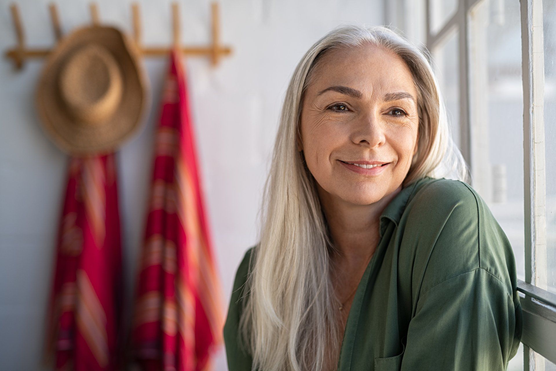 Woman with long gray hair smiles, leaning near a window. Red scarf and hat hang on a rack behind her.