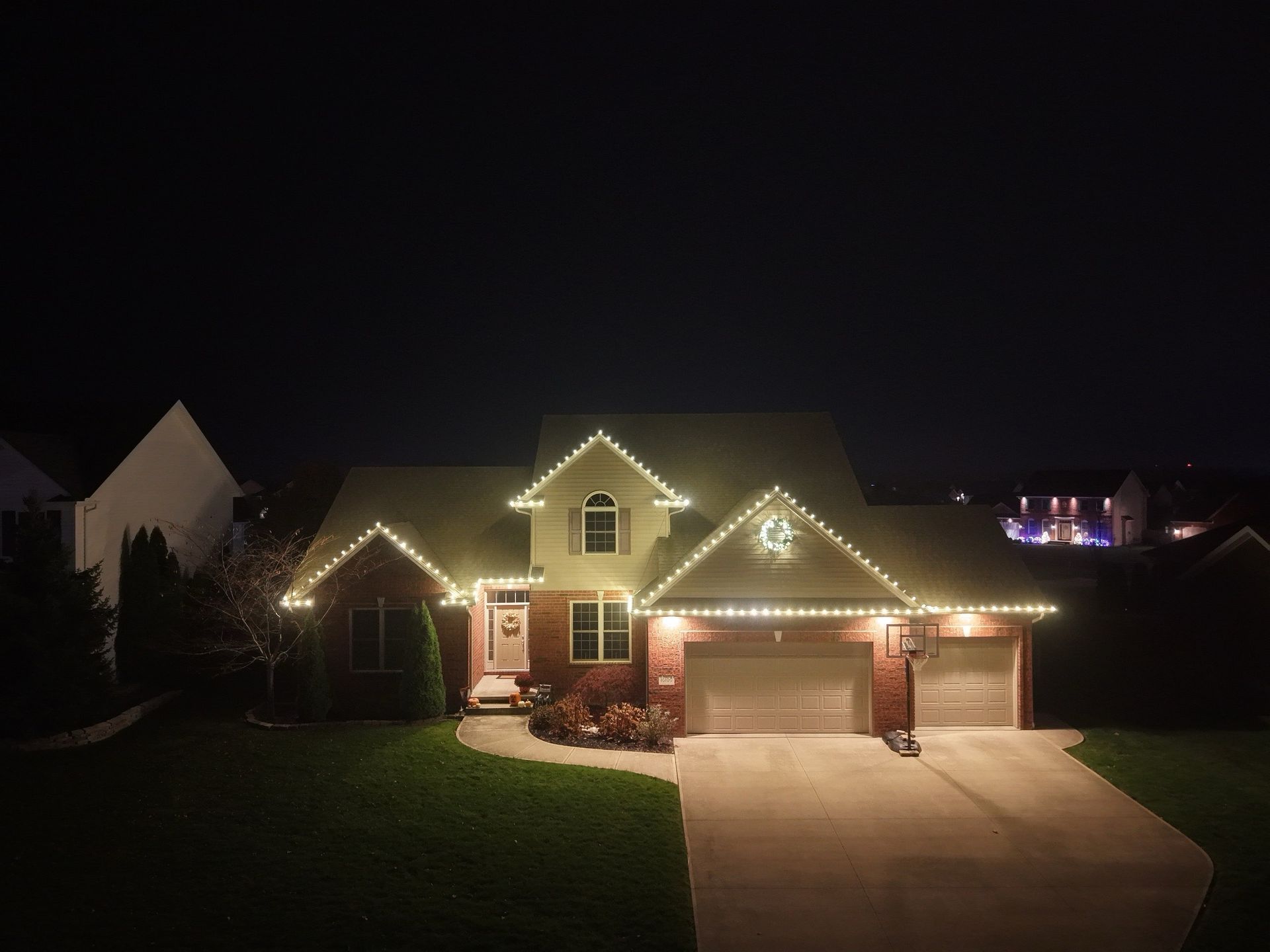 House at night, illuminated with white Christmas lights along the roofline. Garage, lawn, and surrounding homes visible.
