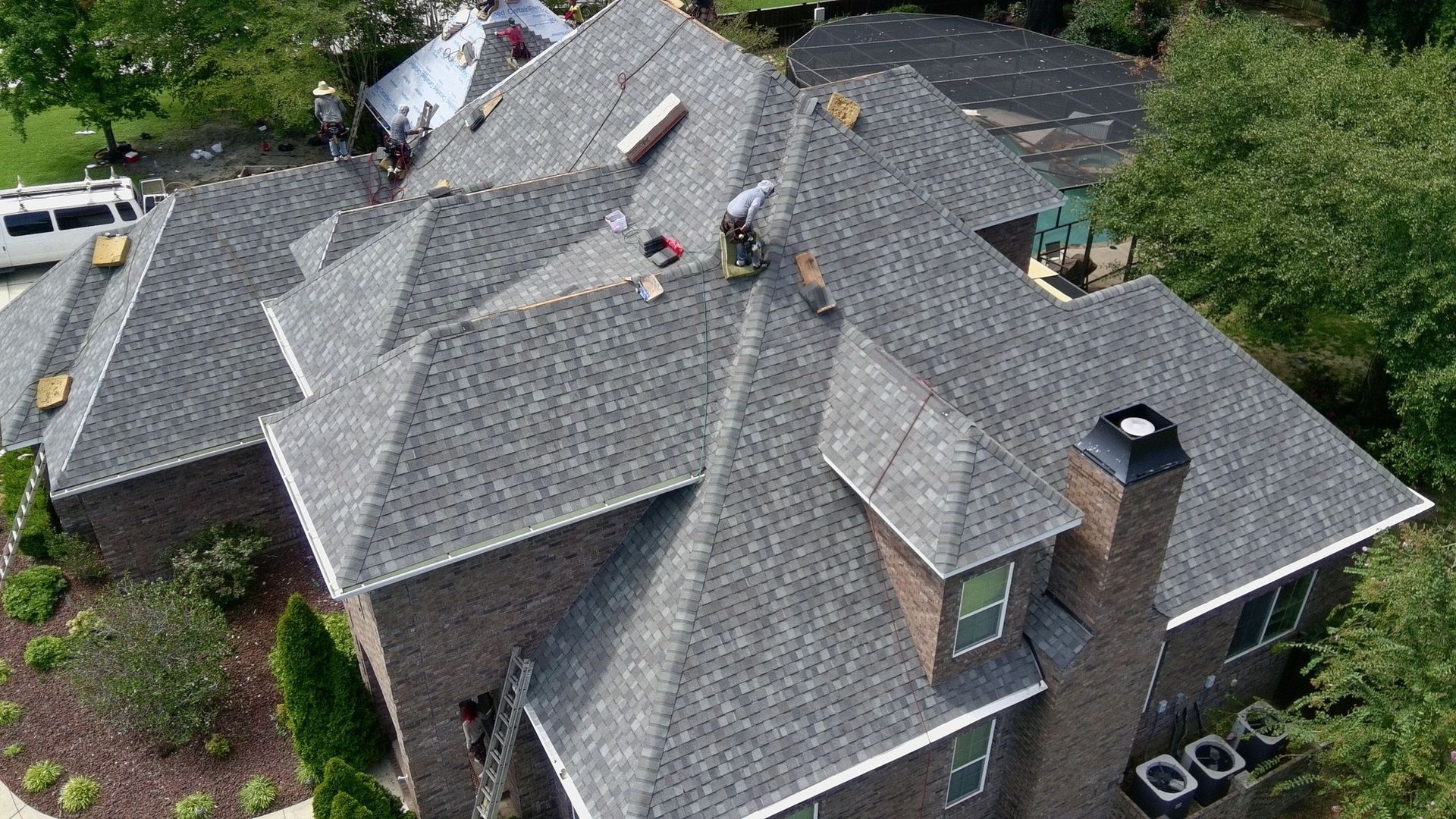 Roofers working on a gray shingle roof of a two-story brick house, with a white van nearby.