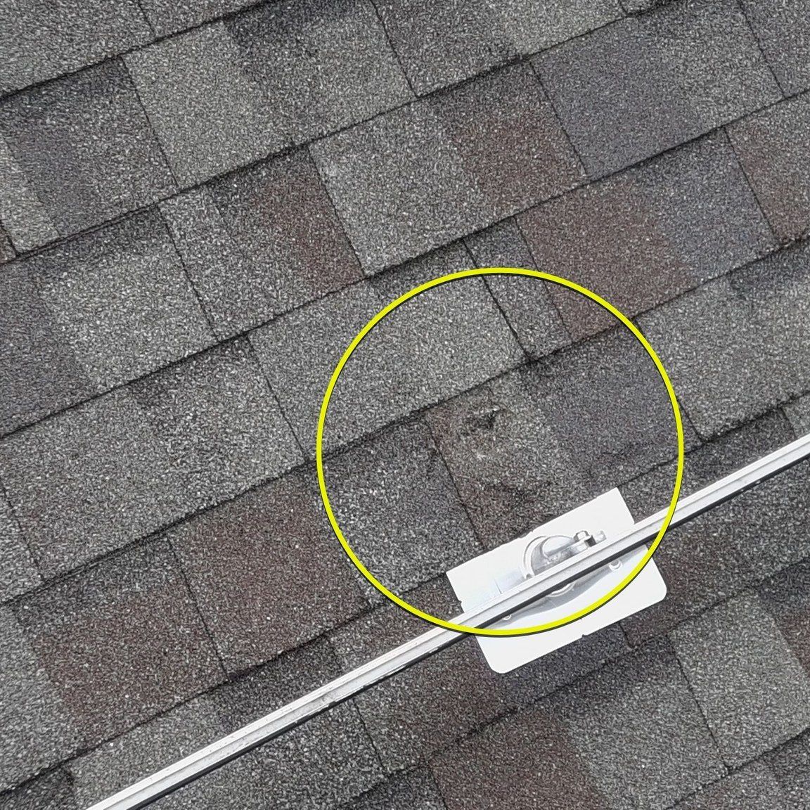 Close-up of a roof with dark gray shingles. A yellow circle highlights a possible leak spot. A solar panel mounting bracket is visible.