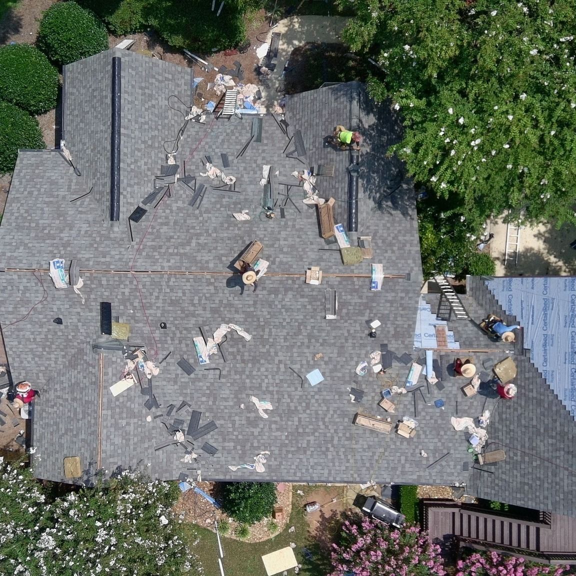 Overhead view of roof repair in progress; debris scattered, workers on roof, surrounded by trees and landscaping.