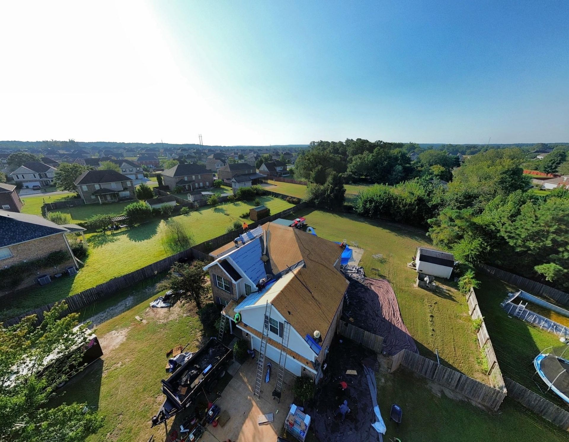 Aerial view of a house with damaged roof and surrounding suburban neighborhood under a blue sky.