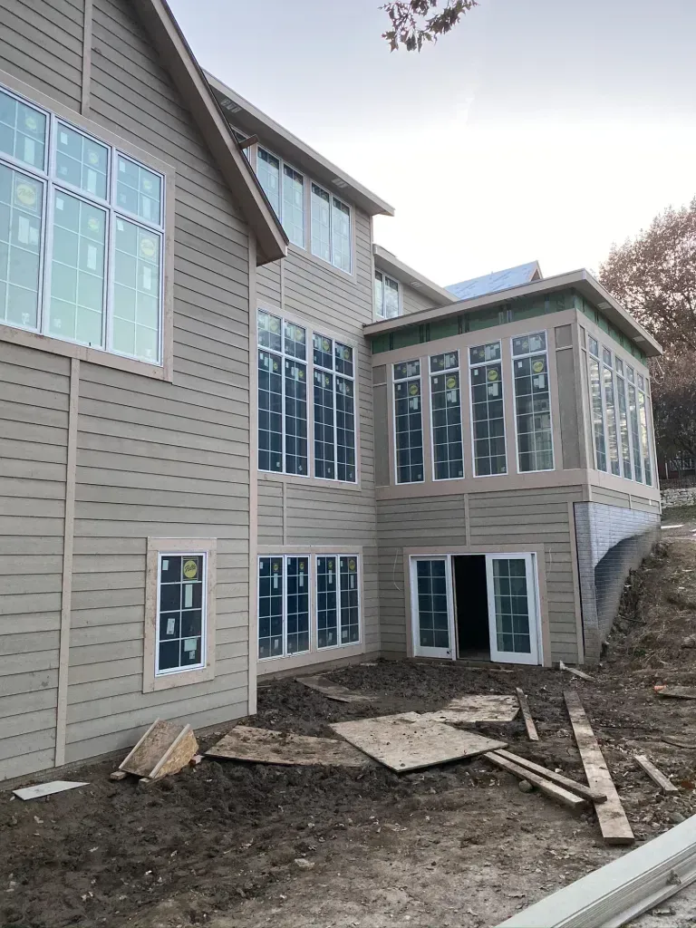 Exterior of a house under construction with many windows and beige siding. Dirt and construction debris are visible.