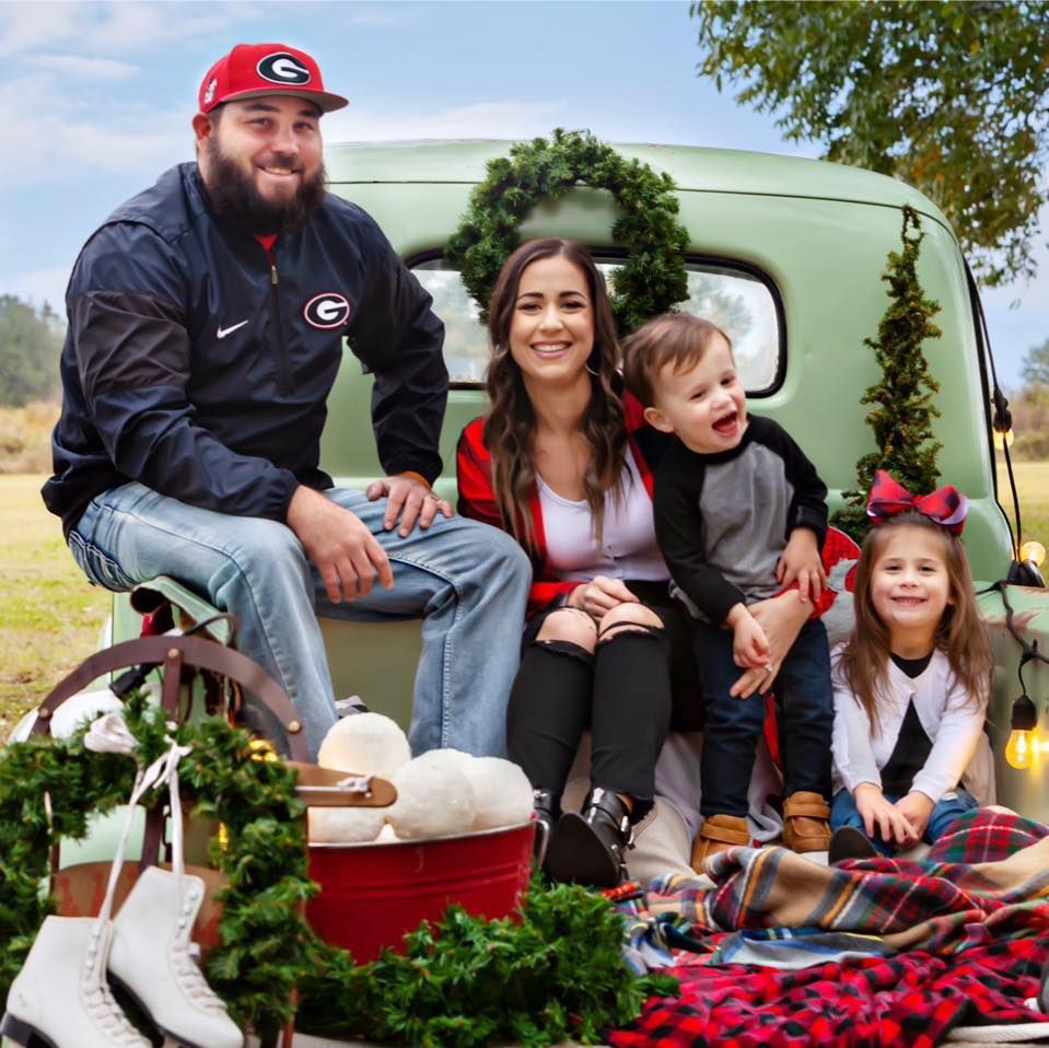 Family poses in a vintage green truck bed decorated for Christmas. Includes wreath, lights, snowballs, and ice skates.