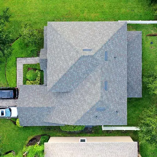 Overhead view of a house with a gray shingled roof, lawn, and adjacent driveway with two parked cars.