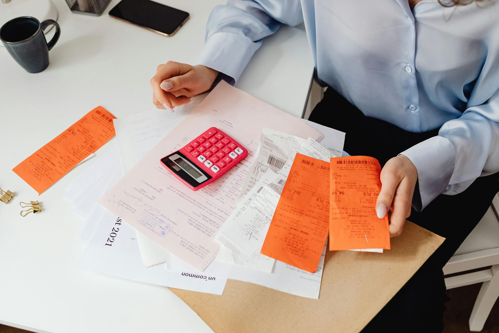 Person in a light blue shirt working at a white desk with a pink calculator and several orange receipts.