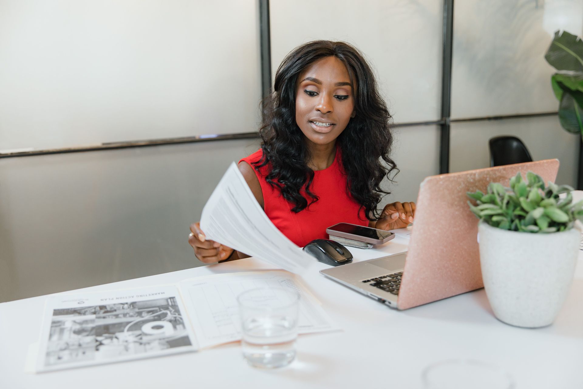 Person working on a laptop at a white desk with papers, a glass of water, and a small potted plant.
