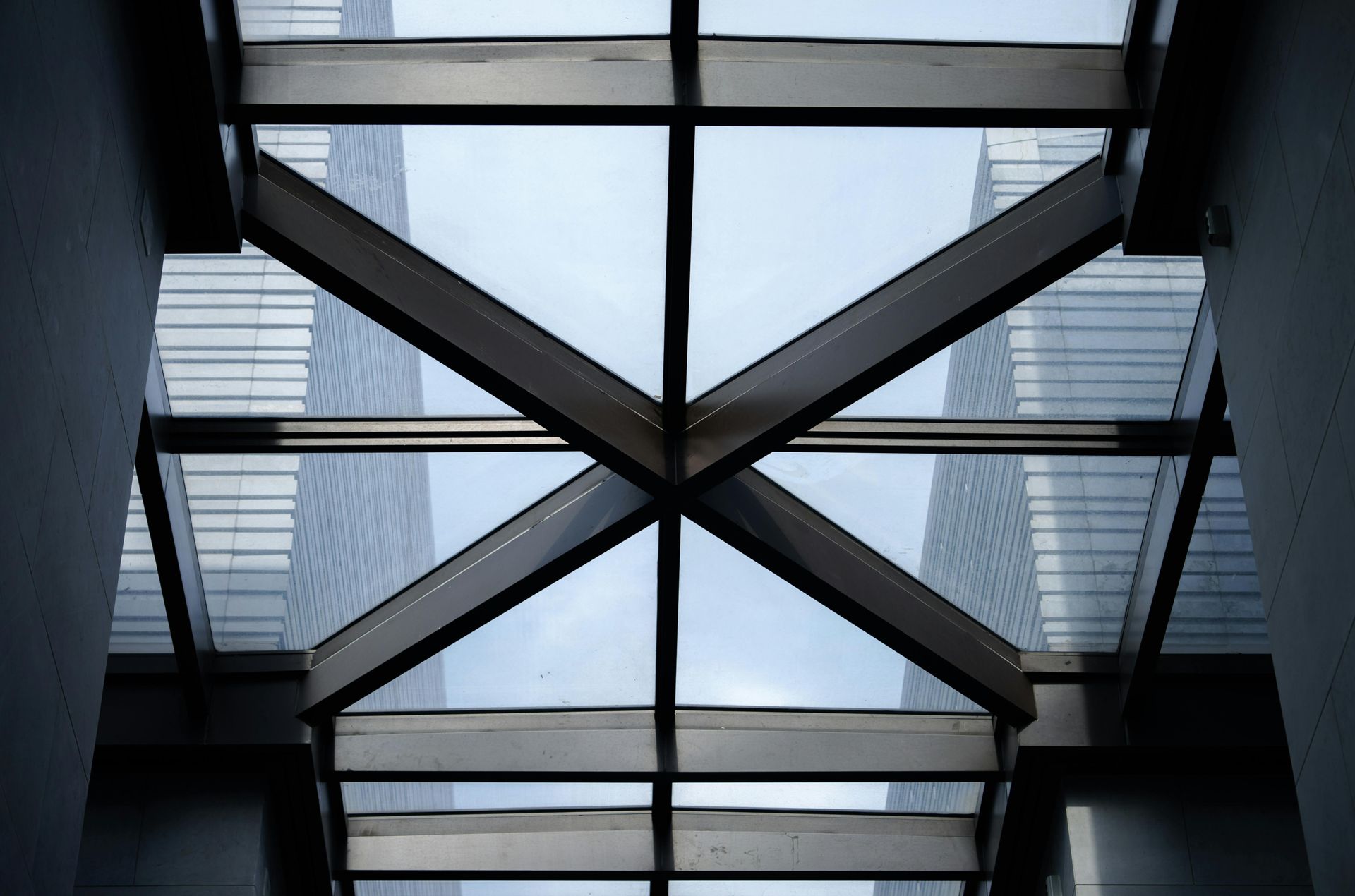 A low-angle view of a glass skylight with dark steel cross-bracing, flanked by tall, modern office building facades.