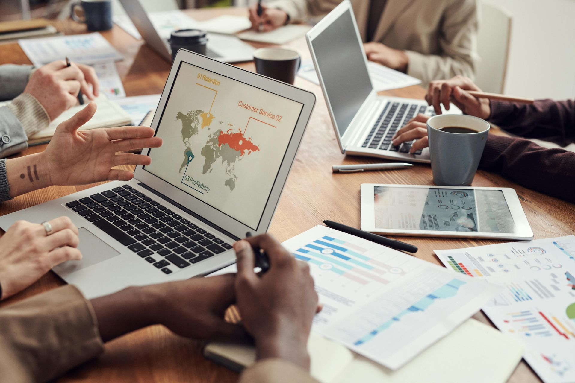 A team collaborating around a wooden table with laptops, charts, and a world map presentation on a screen.