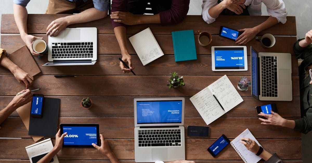 Overhead view of a group collaborating around a wooden meeting table with laptops, tablets, notebooks, and coffee.