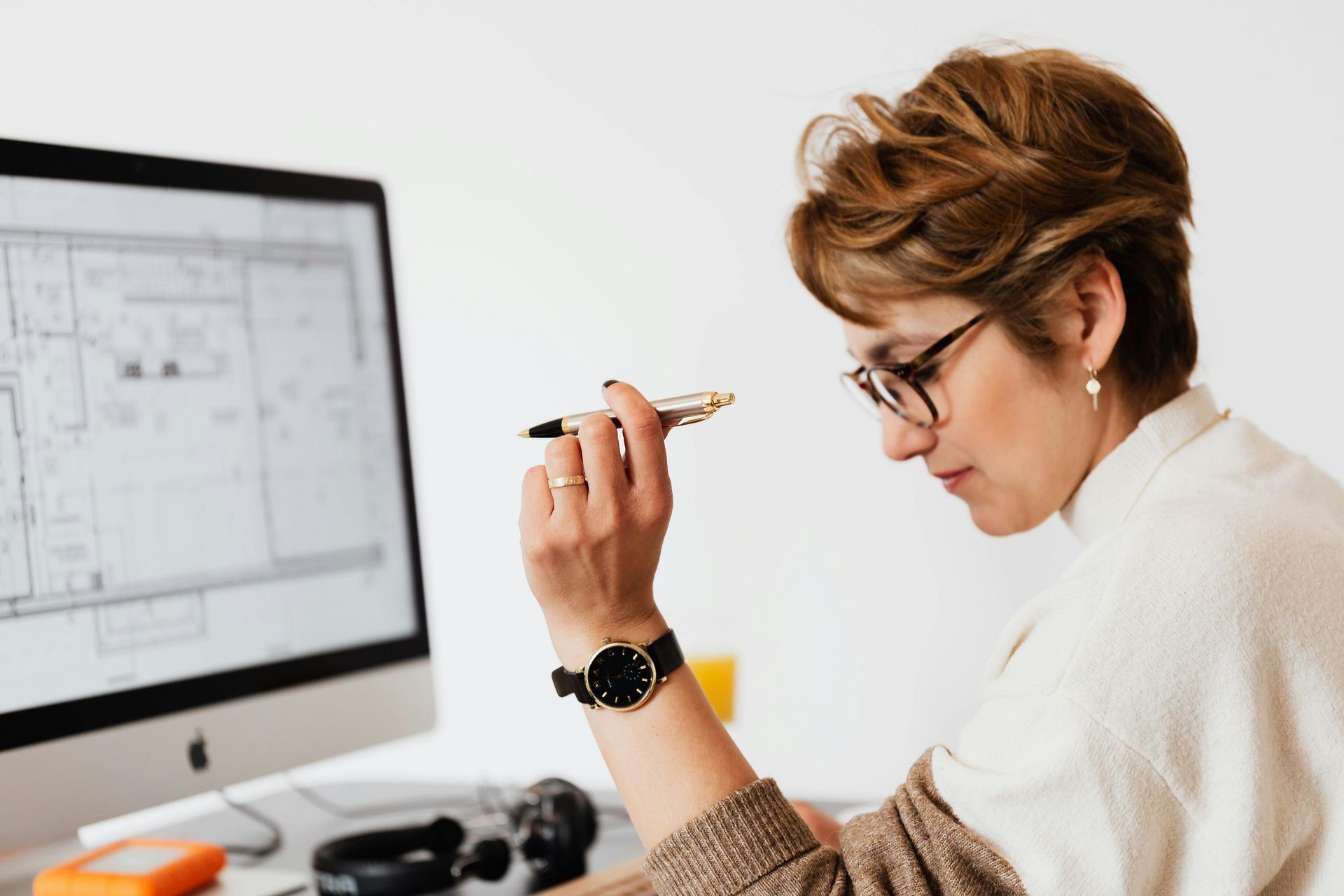 Woman with glasses looks at computer, holding a pen. She wears a watch, working in an office.