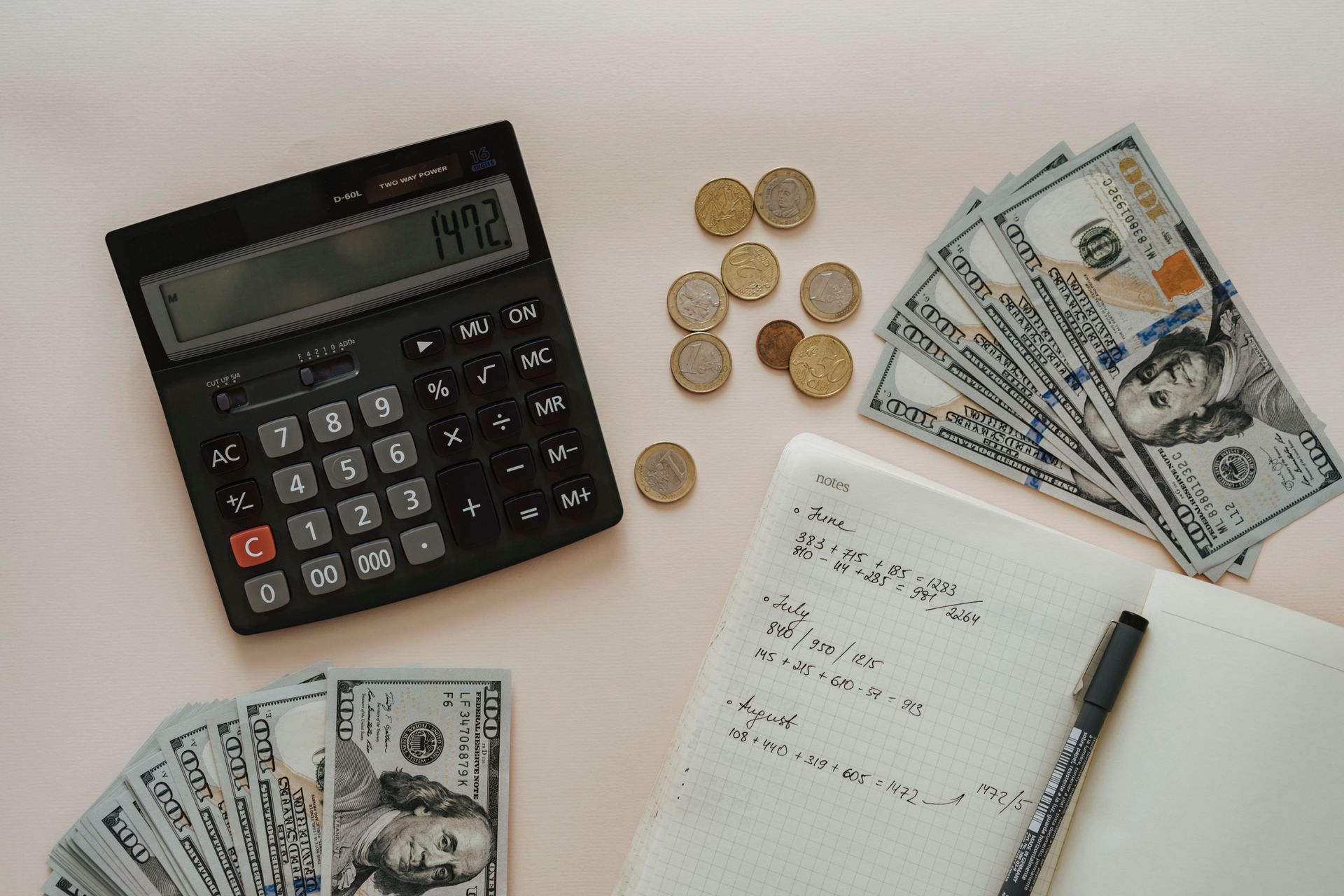 Calculator, coins, dollar bills, and a notepad with pen on a white desk.