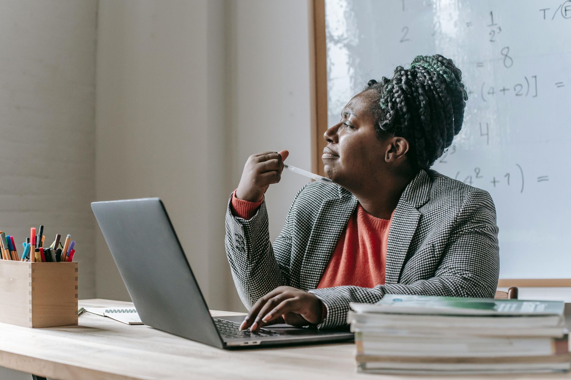A person sitting at a desk with a laptop and books, looking away thoughtfully toward a whiteboard with math equations.