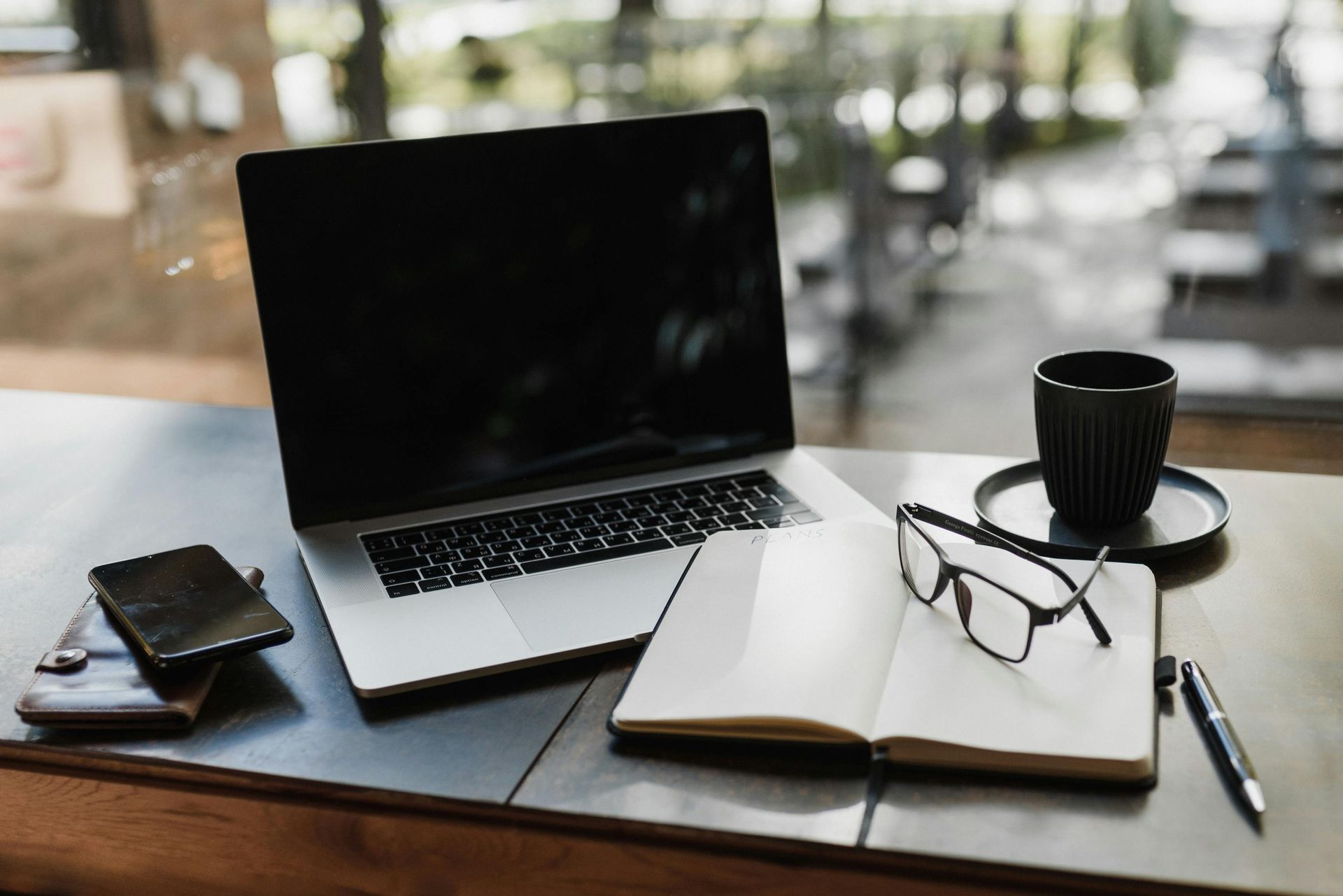 Laptop, open notebook with glasses, phone, and coffee mug on a desk in a blurred interior workspace.
