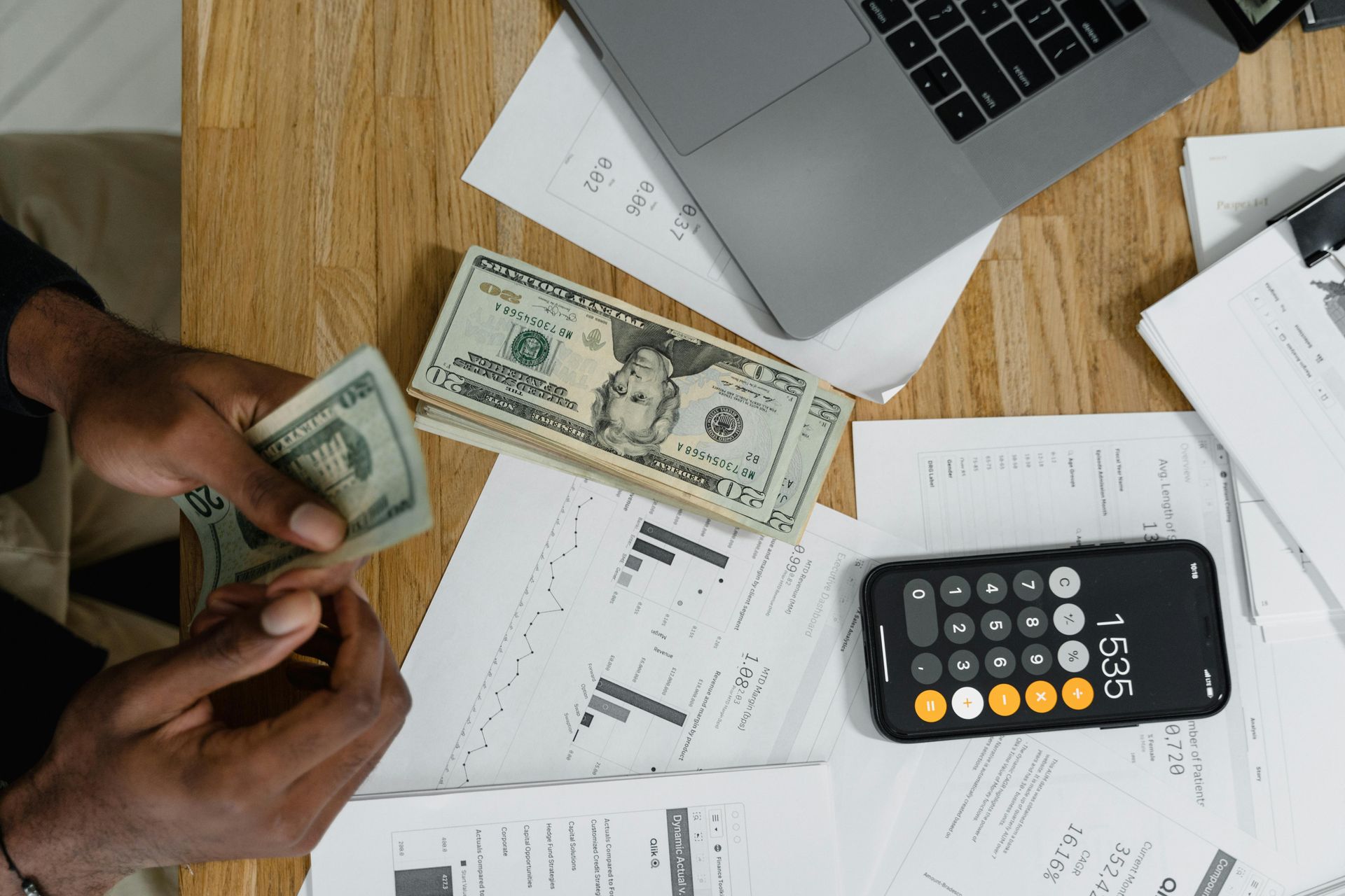 Person counting money on a desk with a laptop, calculator, and documents.