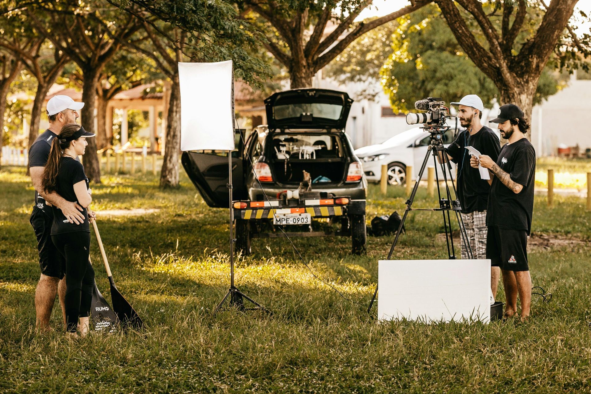 A film crew filming two people with a paddle in a grassy park setting, using cameras, lighting equipment, and a car.