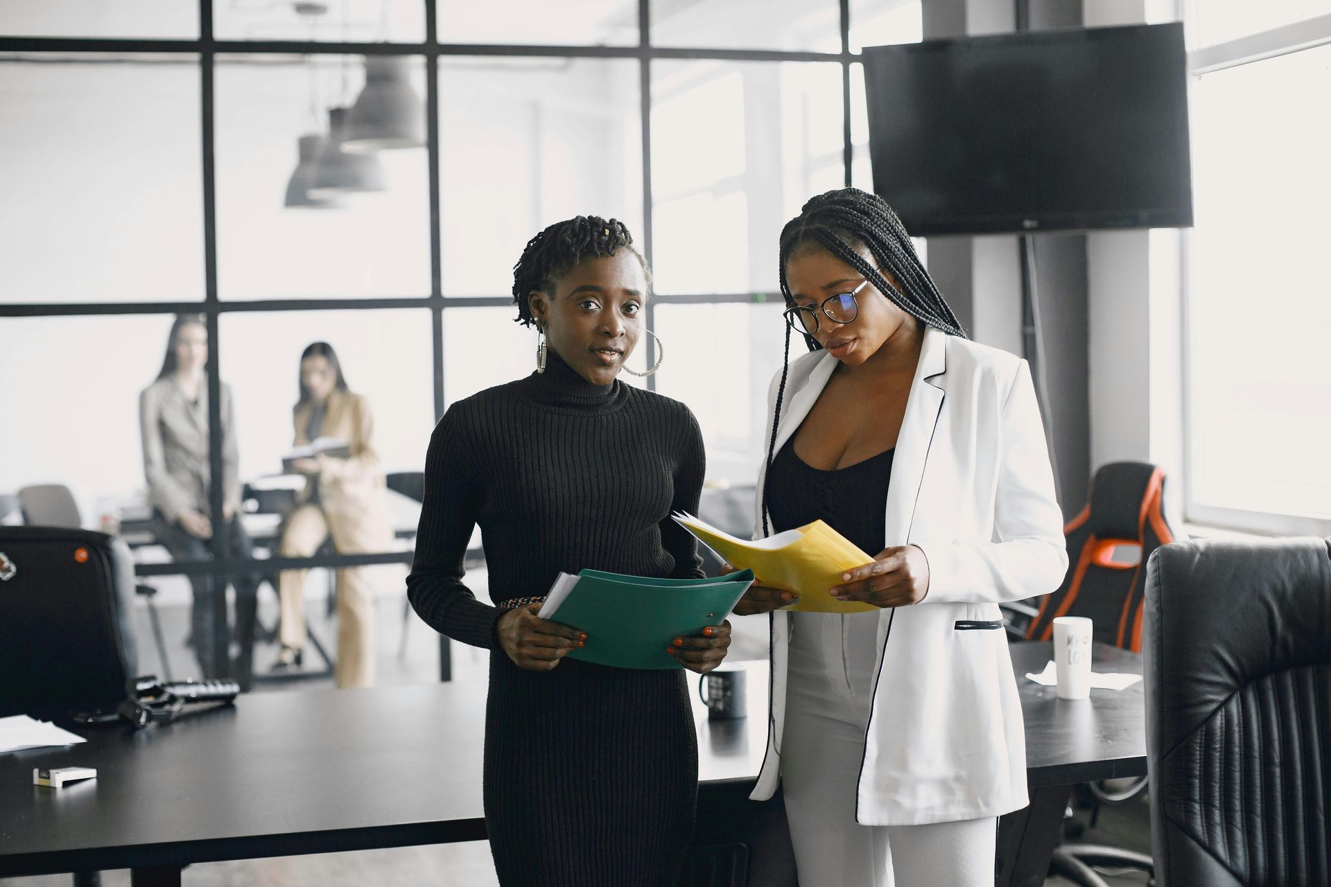 Two professional women standing in a modern office, holding folders and reviewing documents.