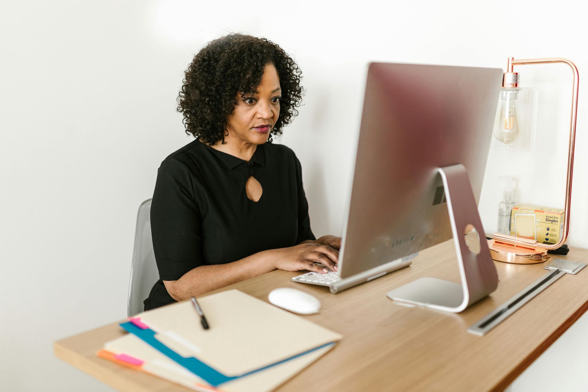 A person with curly hair wearing a black shirt works at a computer on a wooden desk, with a folder and lamp nearby.