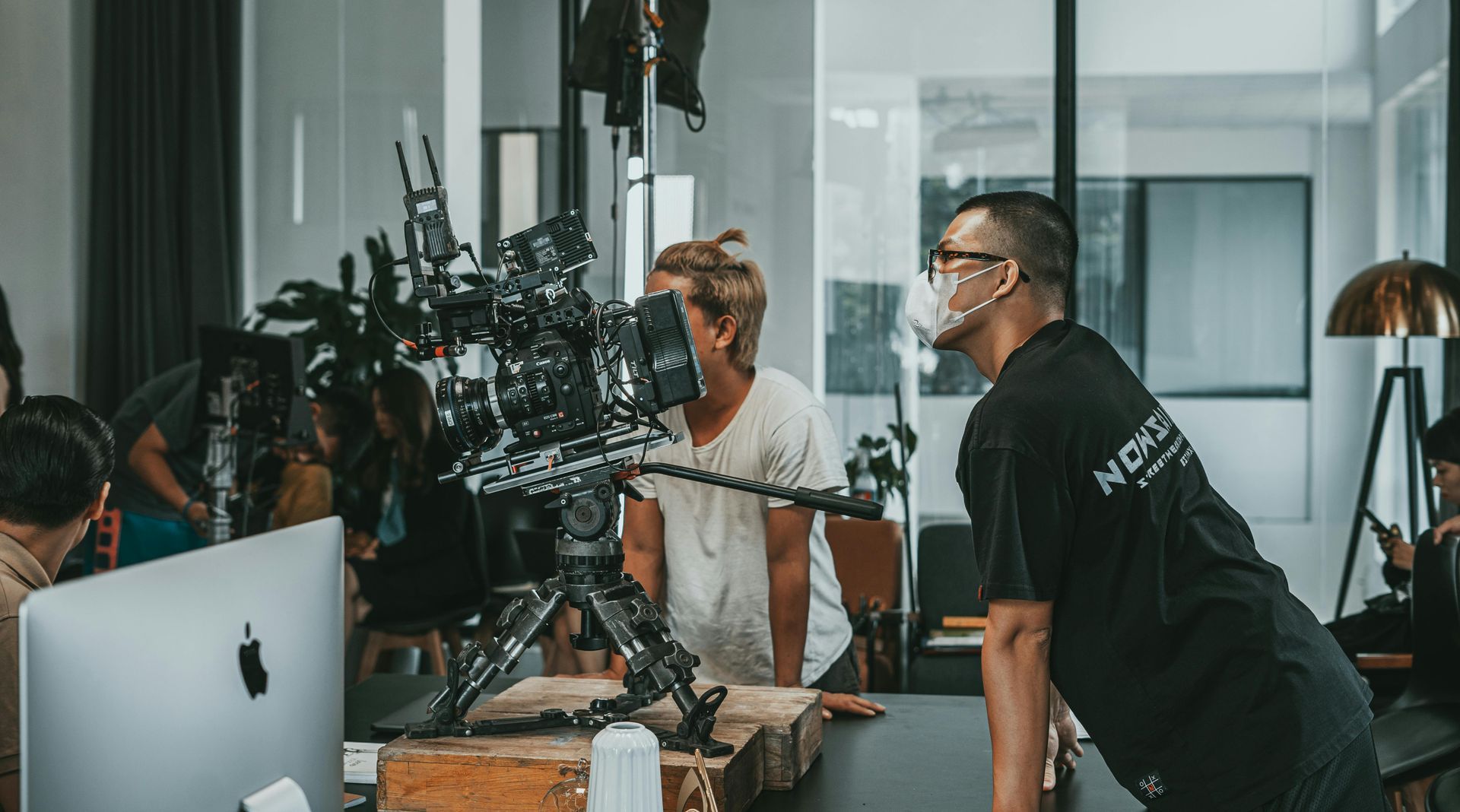 A film crew filming in an office setting with a camera on a tripod, monitored by individuals in casual attire.