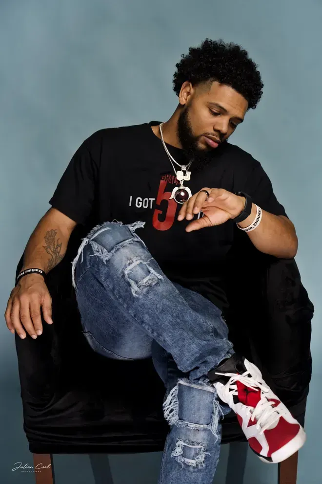 Man seated, looking at watch. Black t-shirt, ripped jeans, white and red sneakers. Blue background.