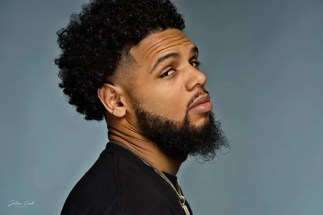 Man with curly dark hair, beard, and sideways gaze against a gray backdrop.