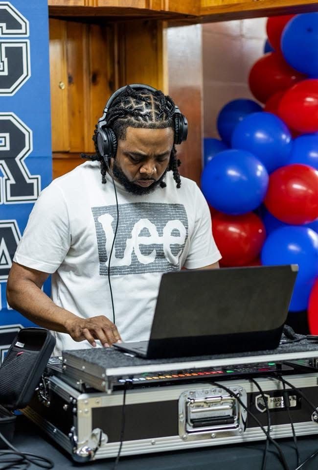 DJ using laptop and mixer with headphones on. Background includes balloons and a sign.