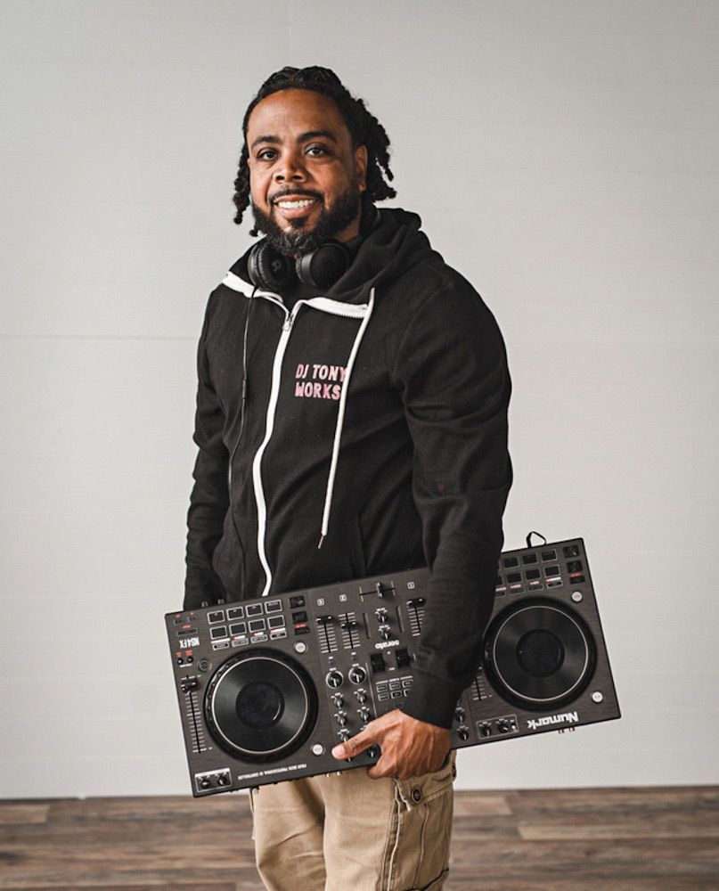DJ holding a sound mixer, wearing a black hoodie and headphones, in front of a white wall.