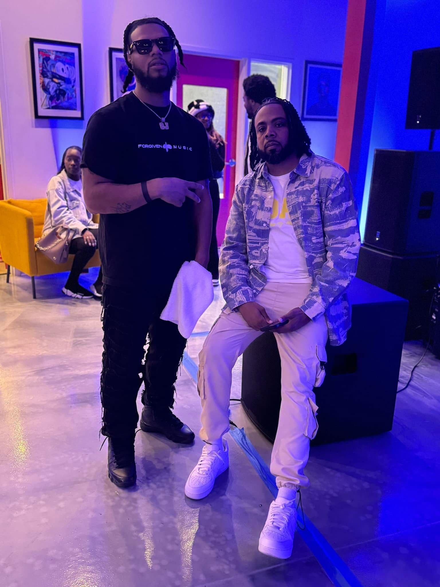 Two men pose inside a room. One stands in black, the other in camouflage and white pants, sitting down.