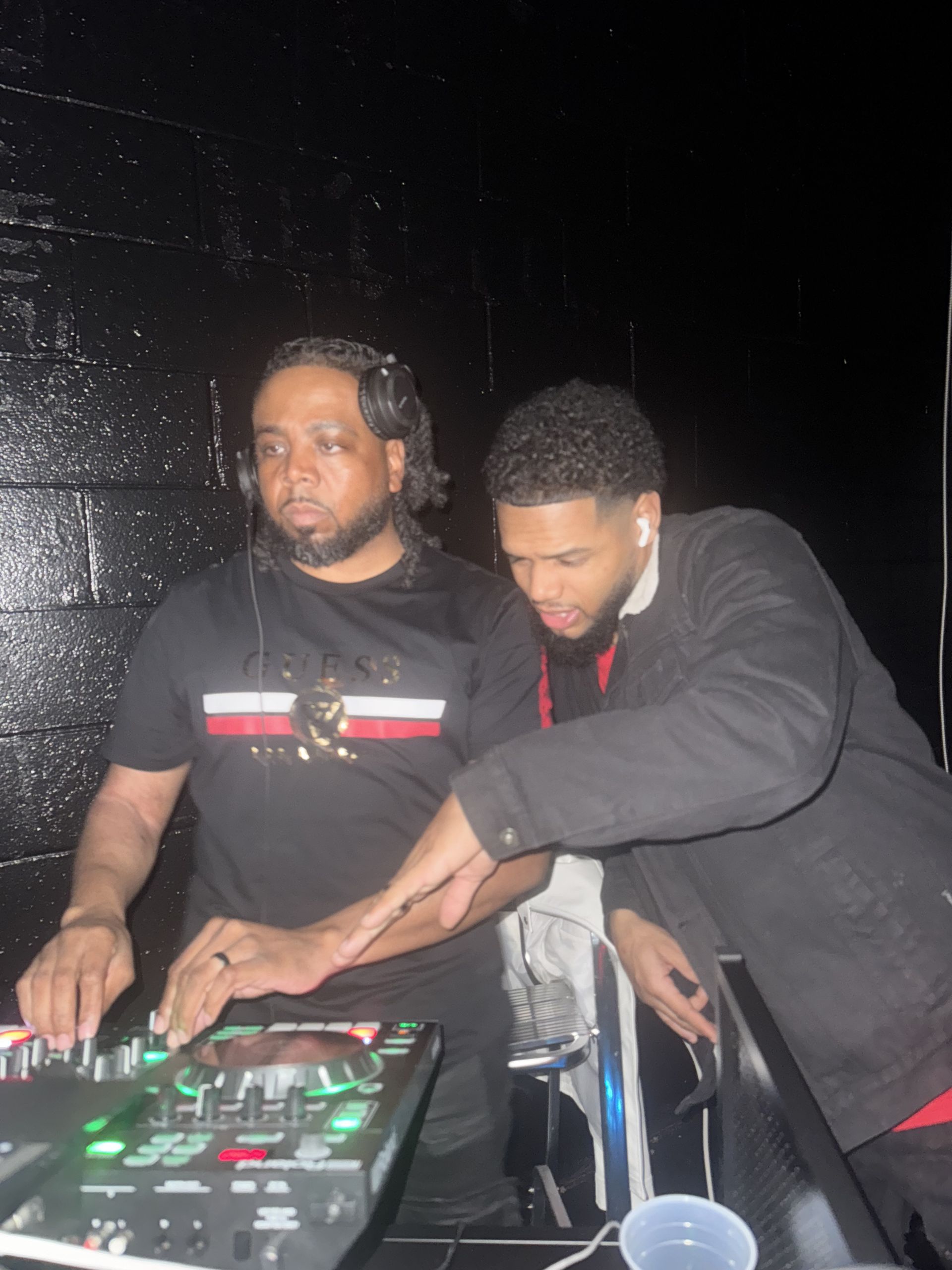 Two men at a DJ setup in a dark setting. One man is wearing headphones, the other appears to be assisting.