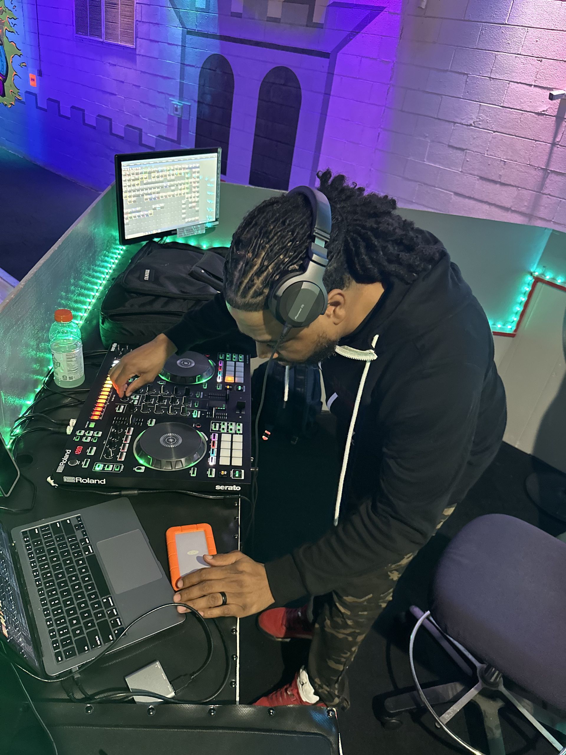 DJ at a console mixing music. They are wearing headphones and a black hoodie. The background is a purple lit party scene.