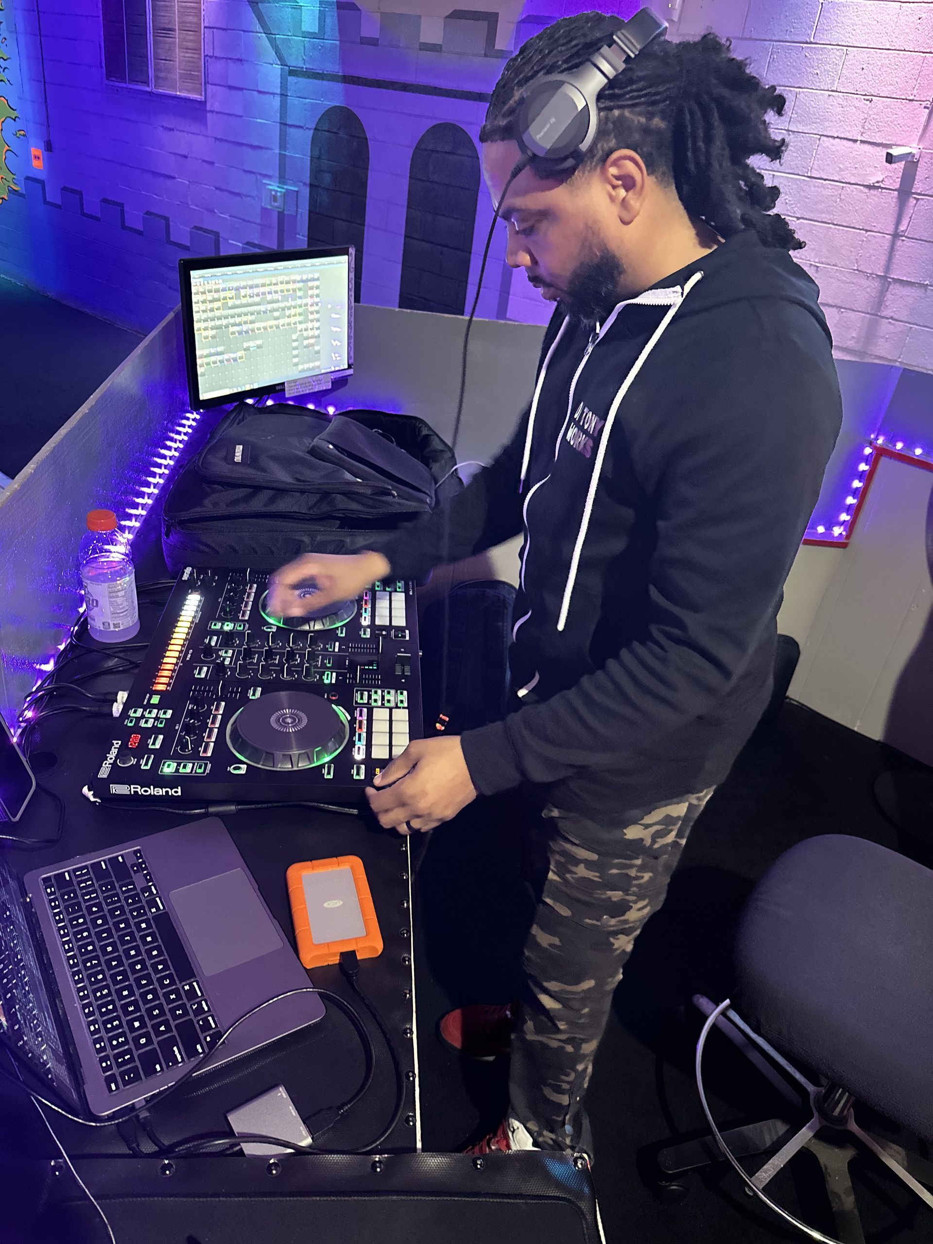 DJ in a dark room with headphones, mixing music on equipment near a laptop and external drive.