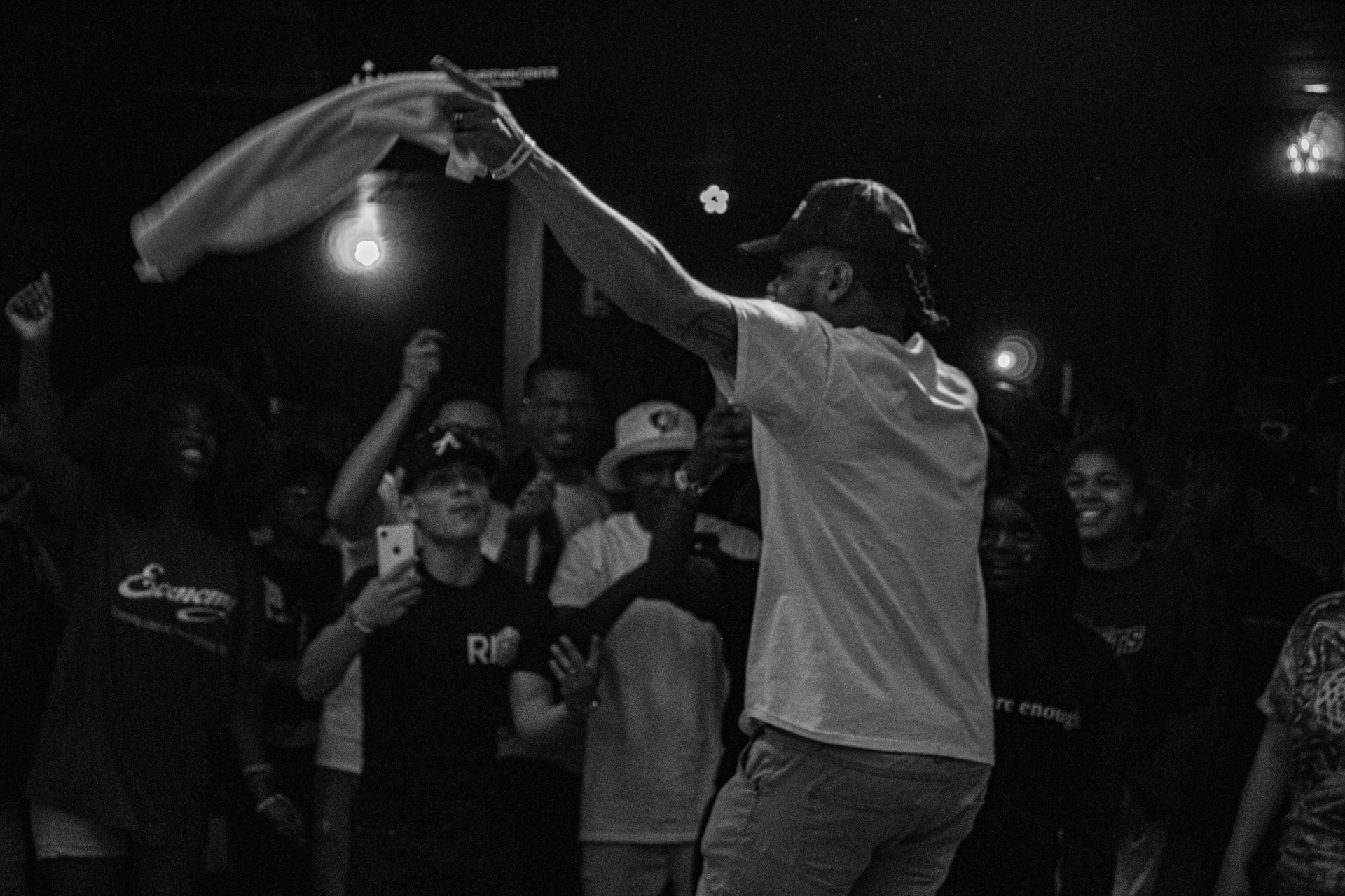 Man waving a cloth above a crowd at night, illuminated by a single light. People cheer and smile.