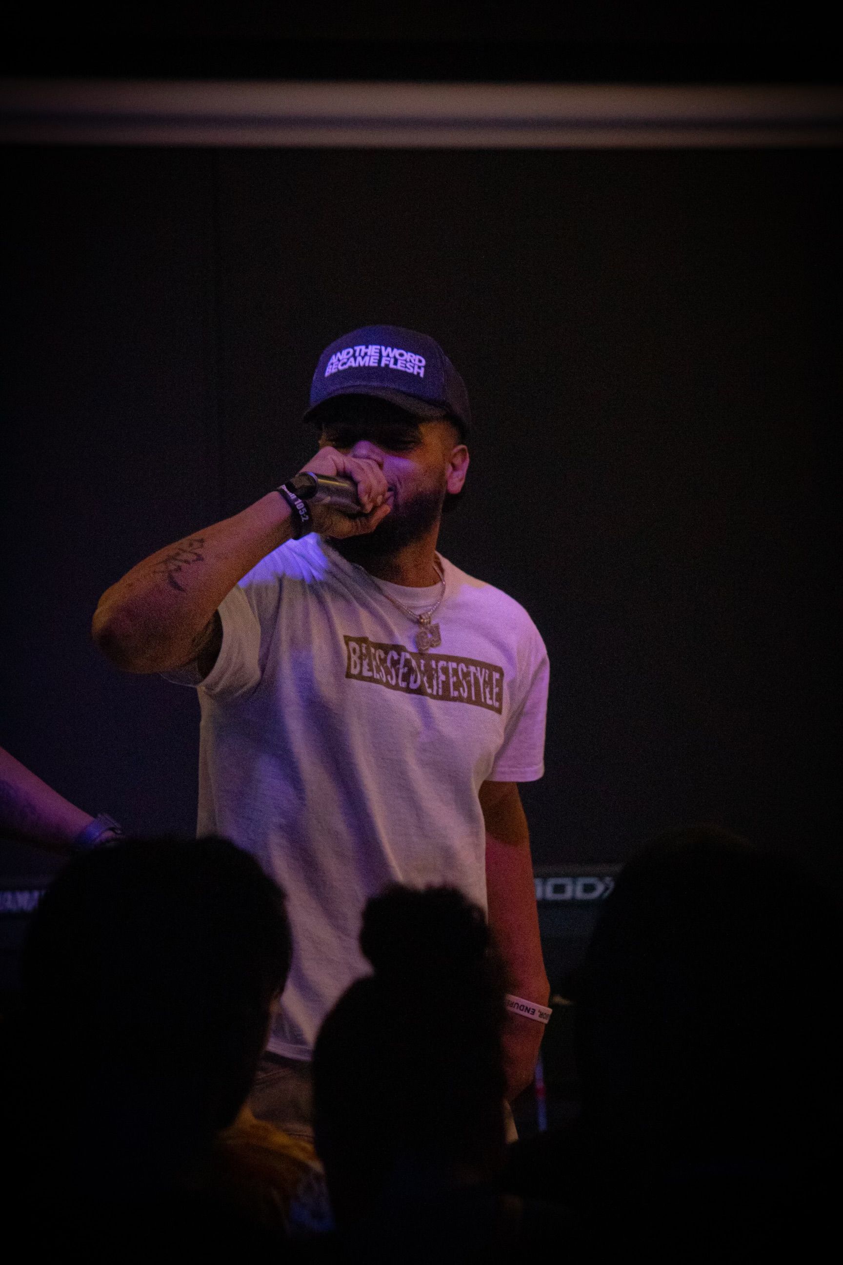 Man wearing a hat and t-shirt, singing into a microphone onstage under dim lighting.