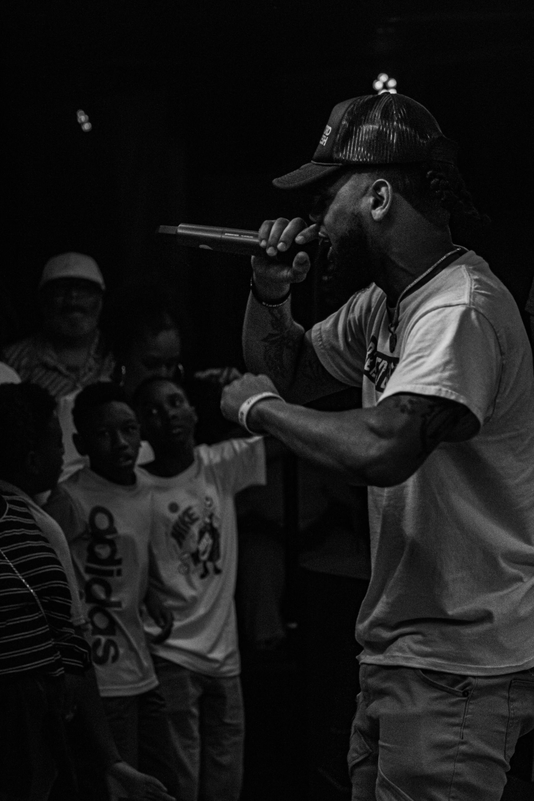 Man rapping on stage, holding a microphone. Crowd watches in the background.