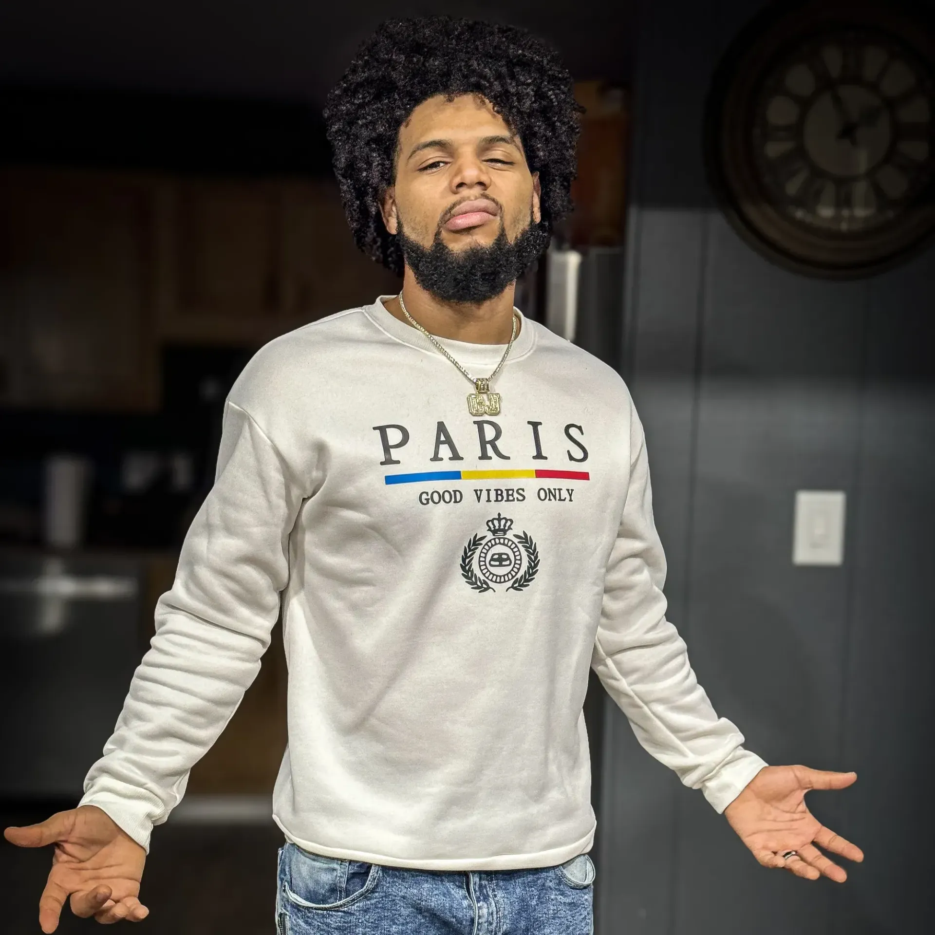 Man with curly hair and beard wearing a Paris sweatshirt, arms outstretched.