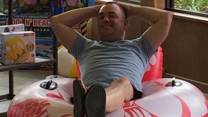 Man relaxing in a red-and-white inflatable ring, leaning back with hands behind his head indoors.