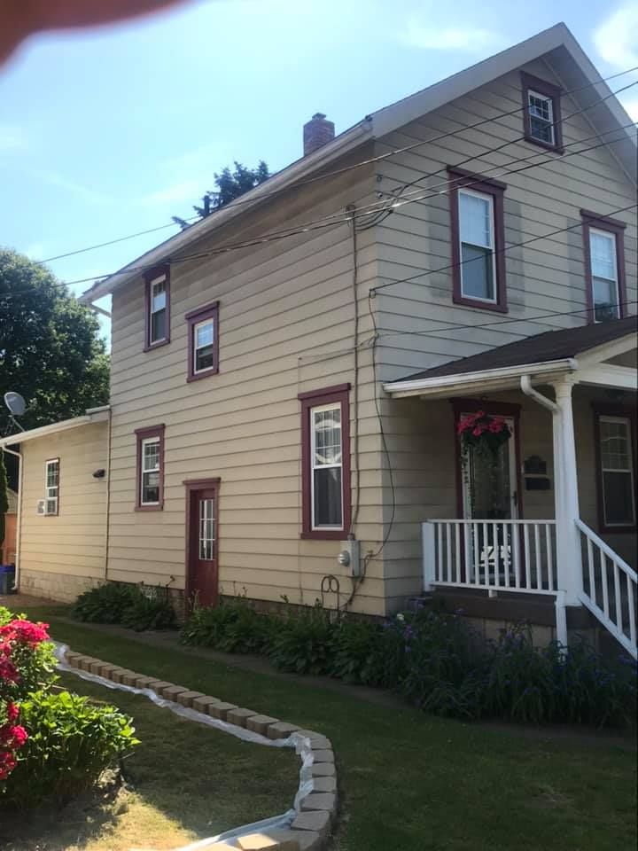 Two-story beige house with red trim around windows and a small porch, with a blue sky background.