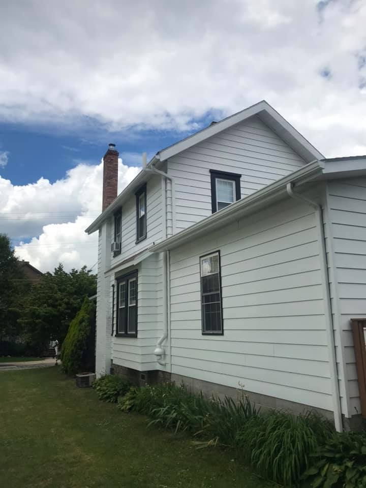 White two-story house with black-framed windows, brick chimney, and green lawn against a cloudy sky.