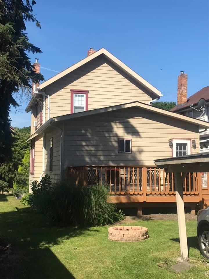 Beige house with a wooden deck, small fire pit on the lawn, and a clear blue sky.