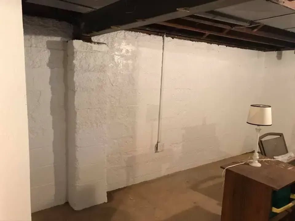 White painted basement walls with a desk, lamp, and chair. Exposed ceiling beams.
