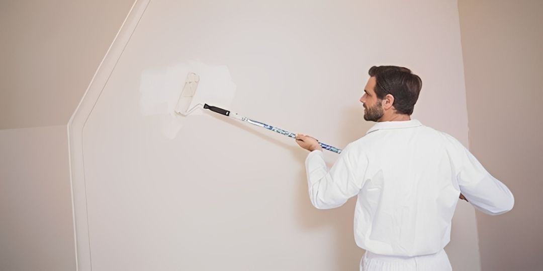 Person in white coveralls paints a wall with a roller. White paint covers the wall.
