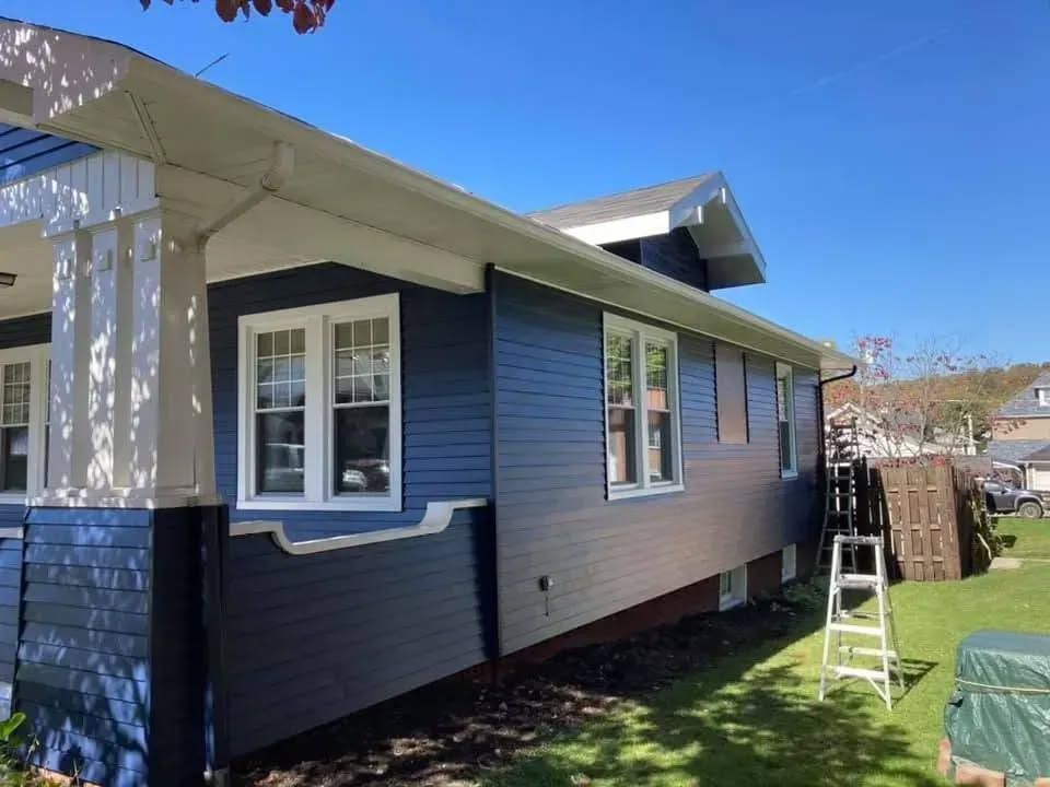 Blue house with white trim, windows, and porch, set against a blue sky, ladder on the side.