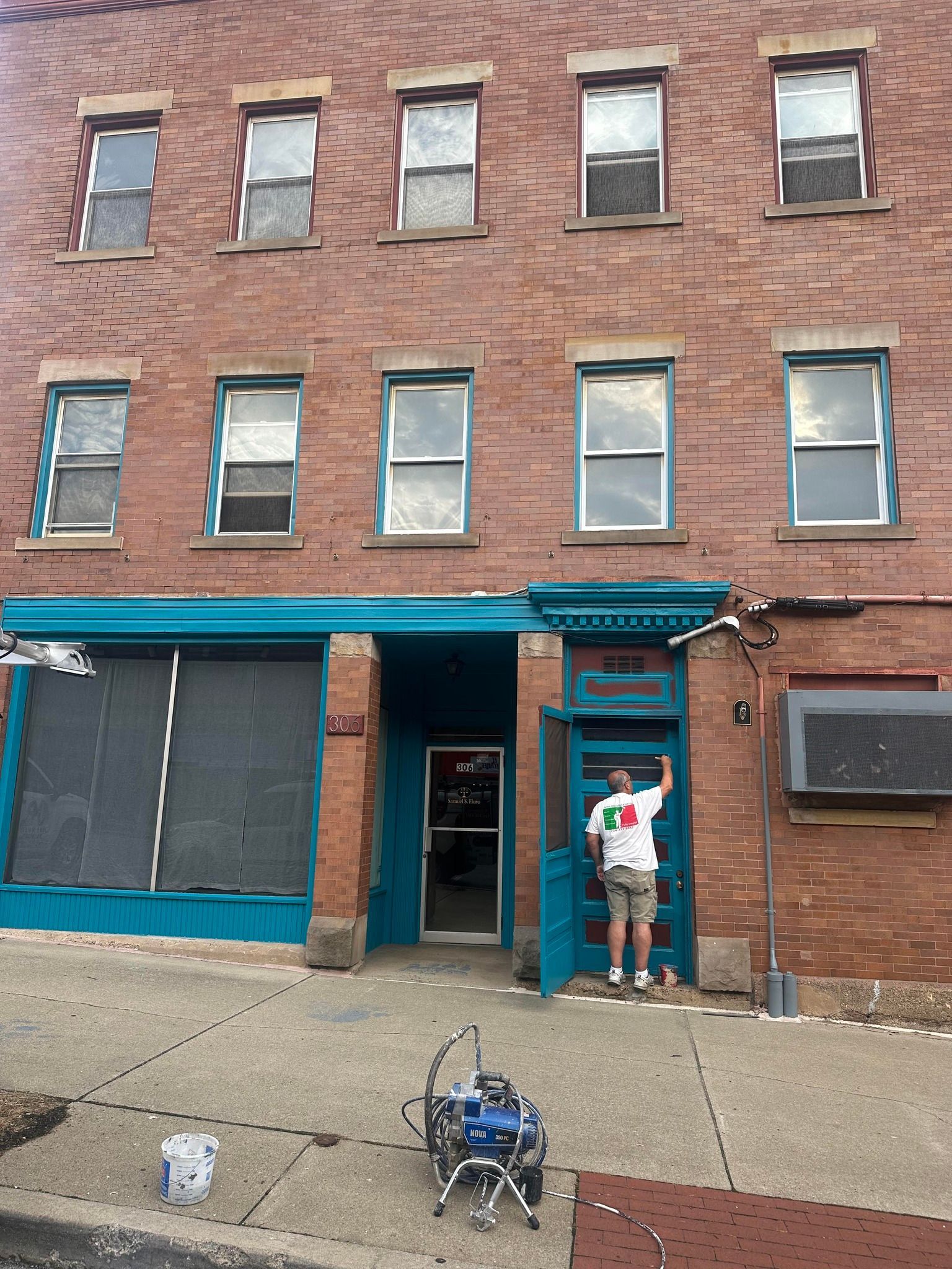 Building with teal door and trim, man painting. Paint sprayer in front.